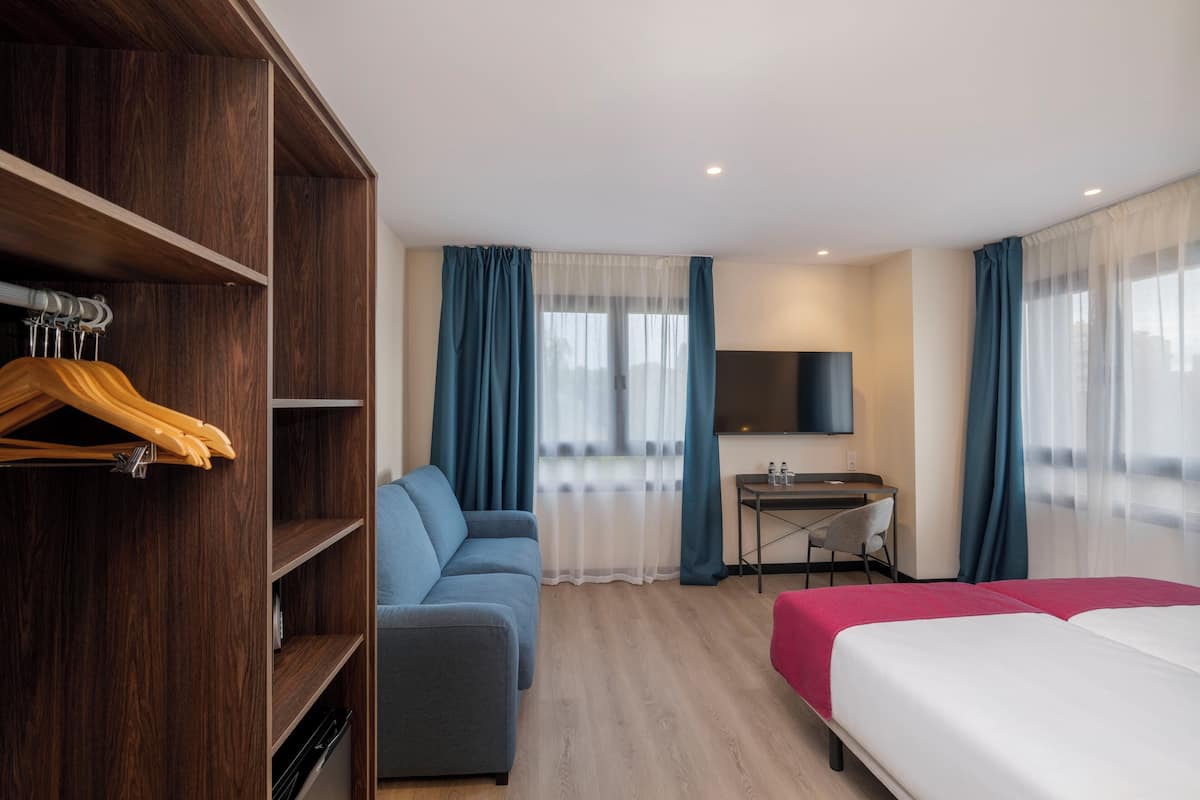 A hotel room with a wooden wardrobe, a sofa, a desk with a chair, a bed, and a TV on the wall.
