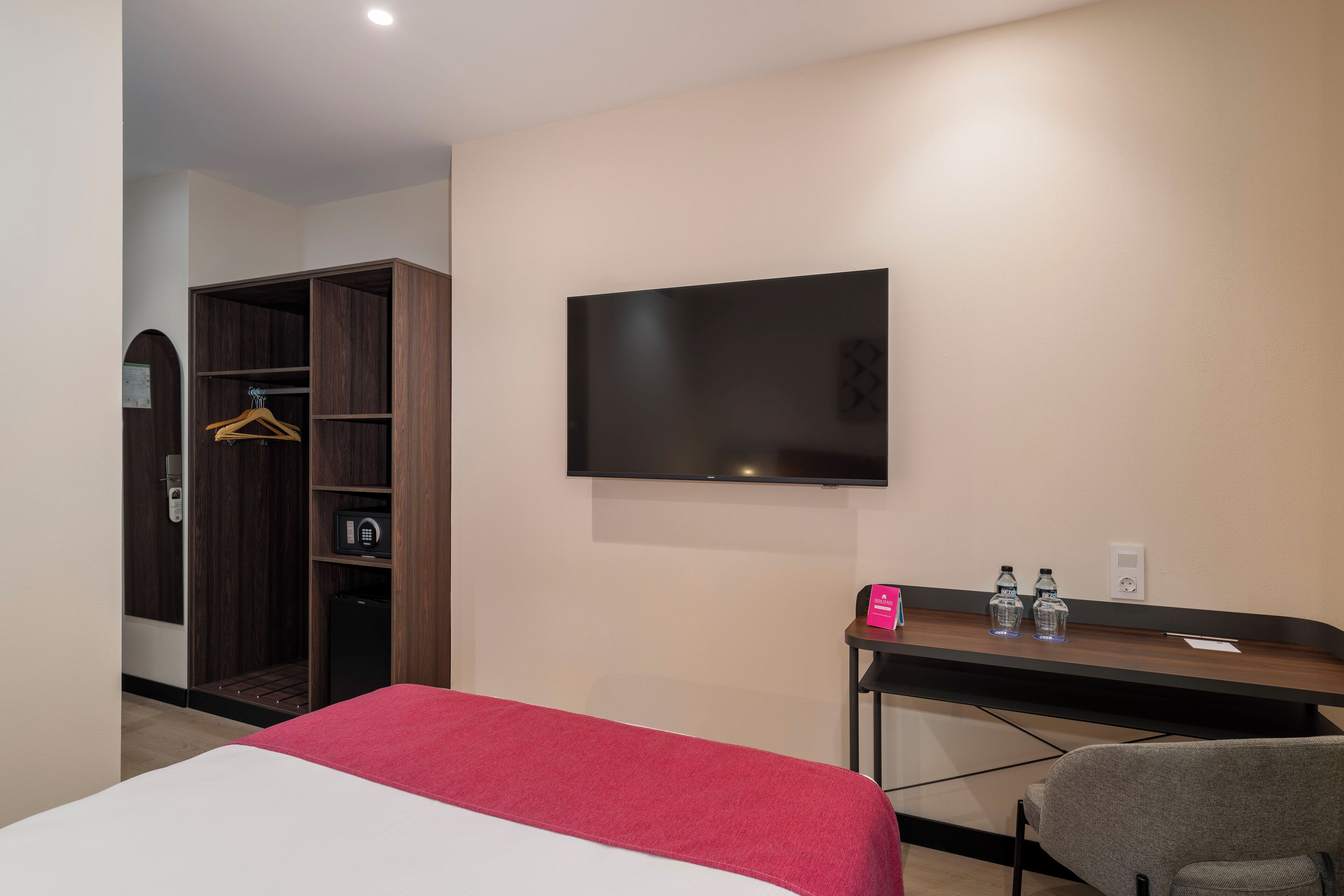 A hotel room with a bed, a TV mounted on the wall, a desk with a chair, and a wardrobe.