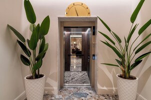 A hallway with a marble floor, a clock on the wall, and two potted plants on either side.