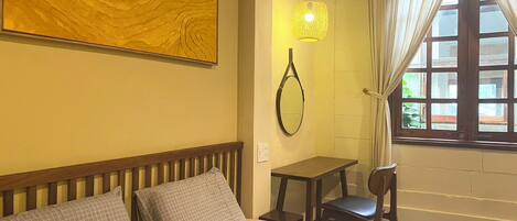 A hotel room with a bed, a desk, a chair, a hanging light fixture, a window with curtains, and a painting on the wall.