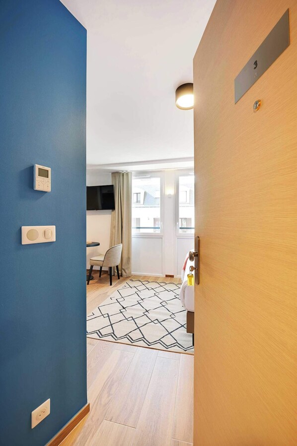 Un couloir avec un mur bleu, une porte en bois et un tapis à motifs menant à une pièce équipée d’un téléviseur et d’un coin repas.