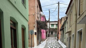 Eine schmale Kopfsteinpflasterstraße, gesäumt von Gebäuden mit traditioneller Architektur, ein Ablassgitter am unteren Rand und ein klarer Himmel darüber.