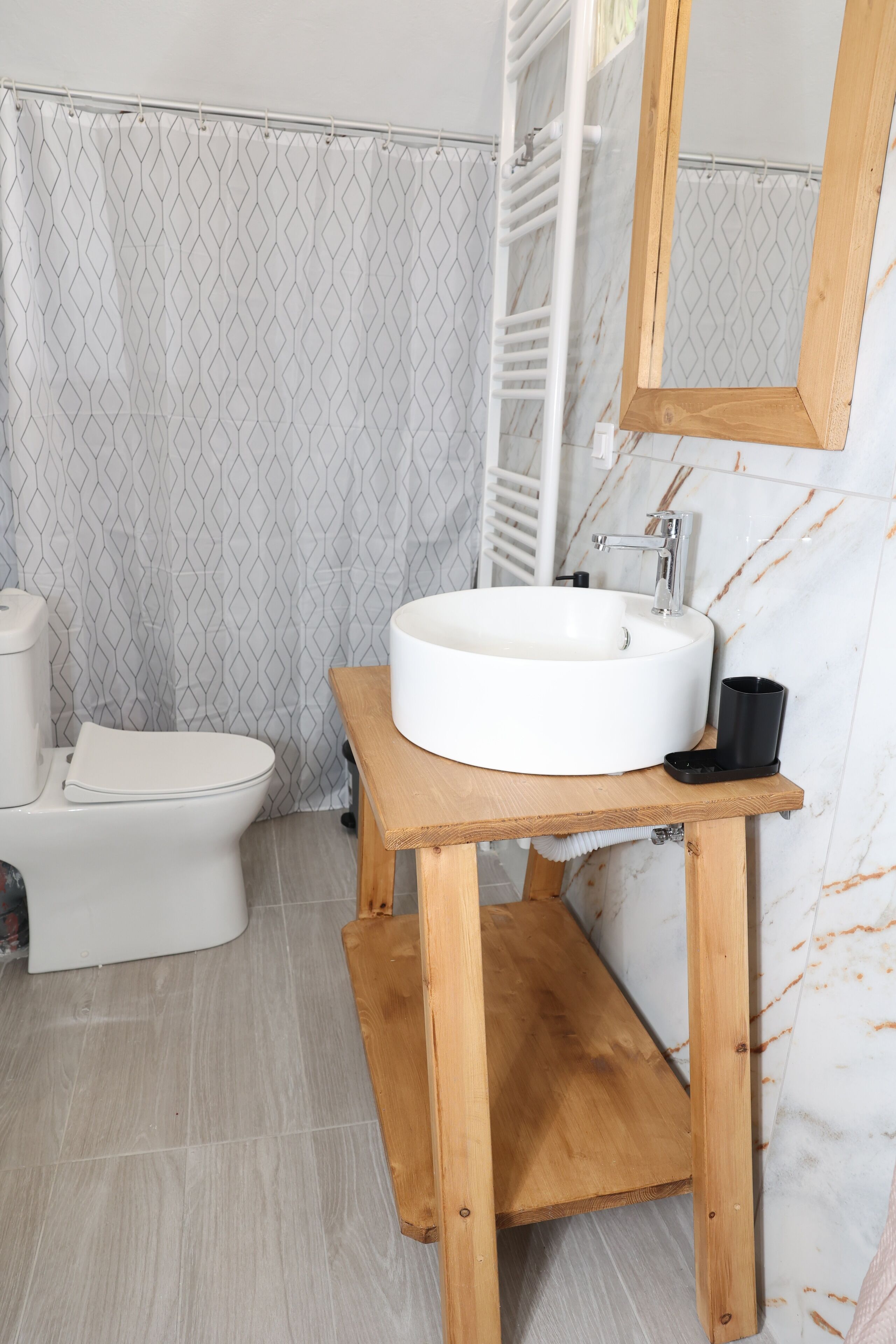 Ein Badezimmer mit weißem Waschbecken, einem Holz-Waschtisch, einer Toilette und einer Duschkabine mit geometrischem Muster.