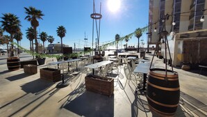 An outdoor seating area with tables and chairs, surrounded by palm trees and a building in the background. - Canet-en-Roussillon: Cozy 1-Bedroom with AC and Parking, Steps from the Beach! (Canet-en-Roussillon)