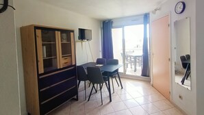 A room with a dining table, chairs, a TV on a stand, a wardrobe, and a sliding glass door leading to an outdoor area. - Canet-en-Roussillon: Cozy 1-Bedroom with AC and Parking, Steps from the Beach! (Canet-en-Roussillon)