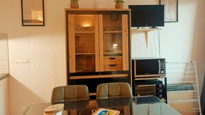 A dining area with a glass table, two chairs, a cabinet with glass doors, a clock, a television, and a microwave. - Canet-en-Roussillon: Cozy 1-Bedroom with AC and Parking, Steps from the Beach! (Canet-en-Roussillon)