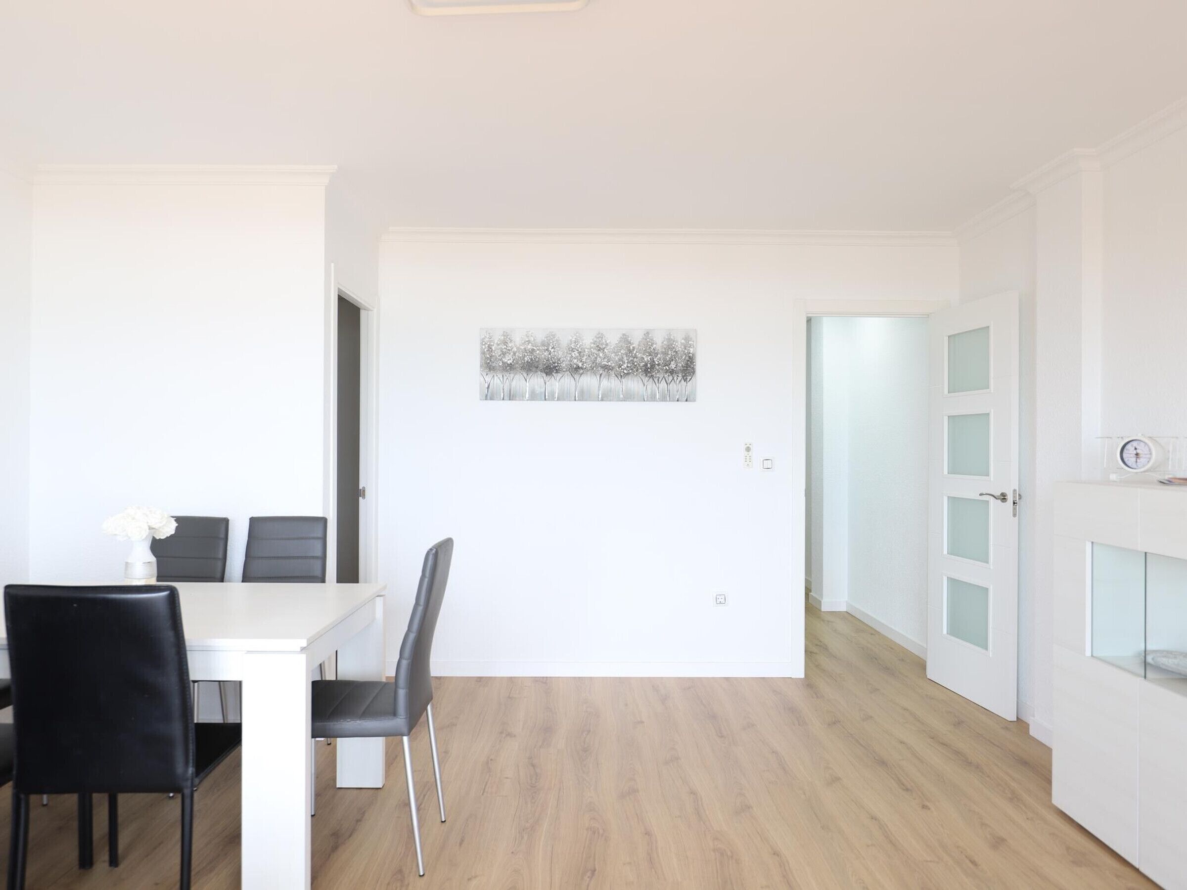Una stanza moderna e minimalista con zona pranzo, caratterizzata da un tavolo bianco e sedie nere. Un quadro appeso al muro, e una porta con pannelli trasparenti.