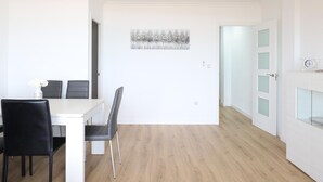 Una habitación moderna y minimalista con zona de comedor que incluye una mesa blanca y sillas negras. Un cuadro cuelga en la pared, y se ve una puerta con paneles de vidrio esmerilado.