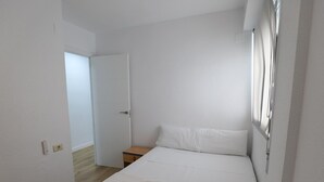 Una habitación limpia y blanca con una cama, una mesita de noche y una puerta que da a otra estancia.