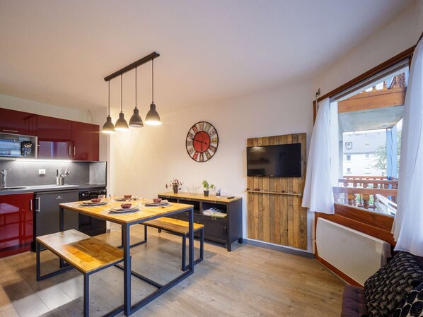A modern kitchen with red cabinets, a wooden dining table with benches, a clock, a TV, and a window with a view of buildings.