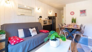 Una sala acogedora con sofá, una mesa de comedor preparada para cuatro personas y una zona de cocina con refrigerador y armarios.