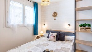Un dormitorio con cama, lámpara de noche, luz empotrada en la pared, ventana con cortinas, un adorno en la pared y una repisa con una planta.