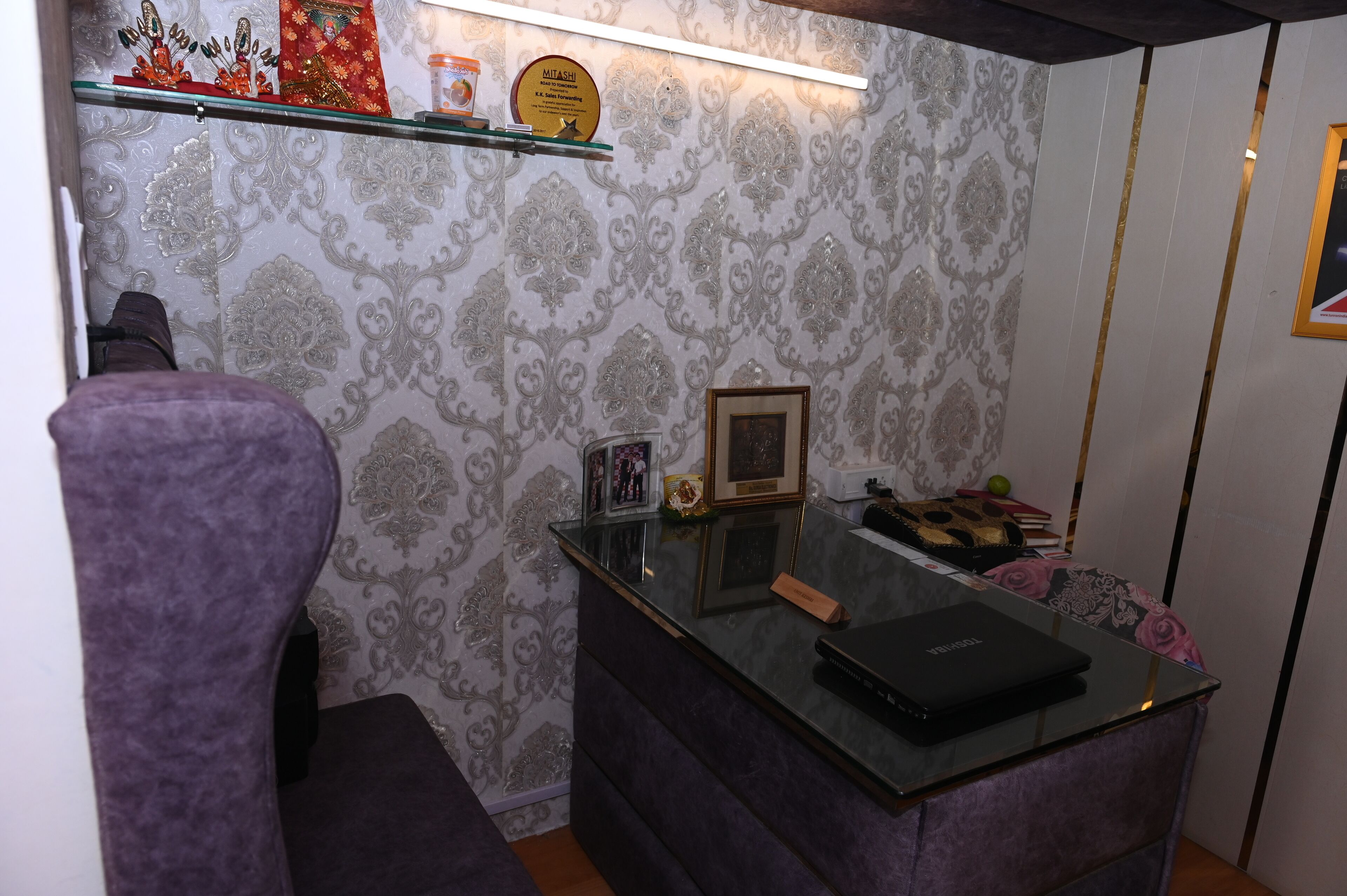 A room with a purple armchair, a glass-top desk with a laptop, a framed picture, and a decorative shelf with items.