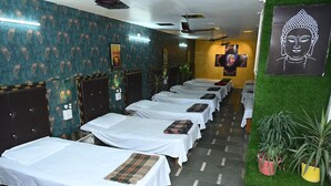 A room with multiple beds aligned in a row, each with a white bedspread and a plaid blanket. There are potted plants on the right side. - Siddhivinayak Temple Guest House (Varanasi)