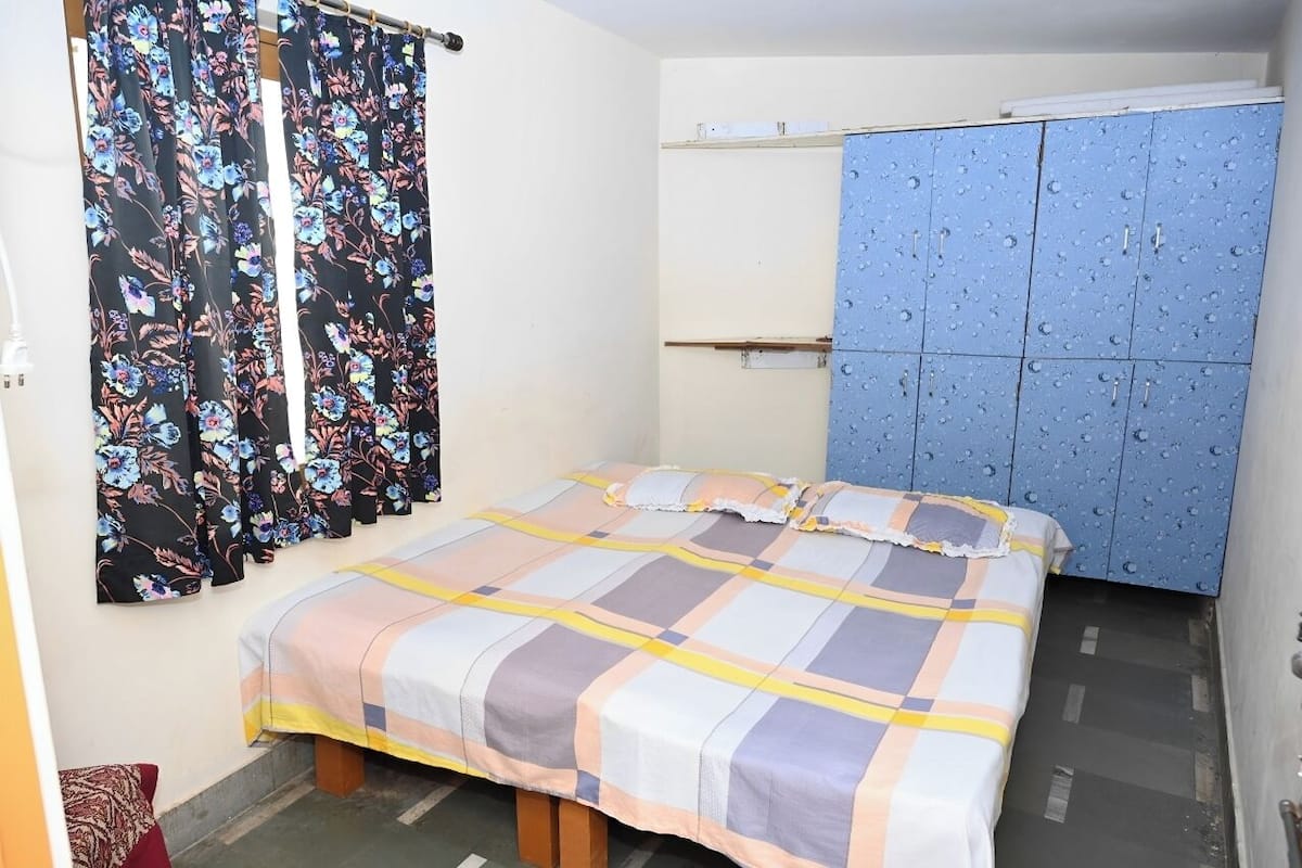 A small, neatly arranged bedroom with a bed, a wardrobe, and a window with floral curtains.