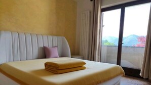 A bed with a yellow blanket and a folded towel, a white headboard, and a view of mountains through a glass door.