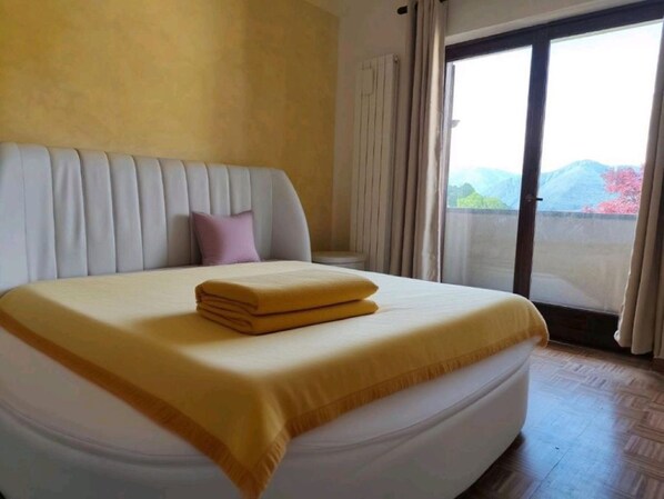 A bed with a yellow blanket and a folded towel, a white headboard, and a view of mountains through a glass door.