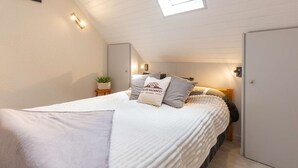 A cozy bedroom with a sloped ceiling, a skylight, a bed with pillows, a bedside table with a plant, and a door.