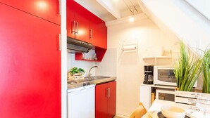 A compact kitchen with red cabinets, a white countertop, a stove, and a microwave. There is a dining area with a table set for two, a chair, and a potted plant.