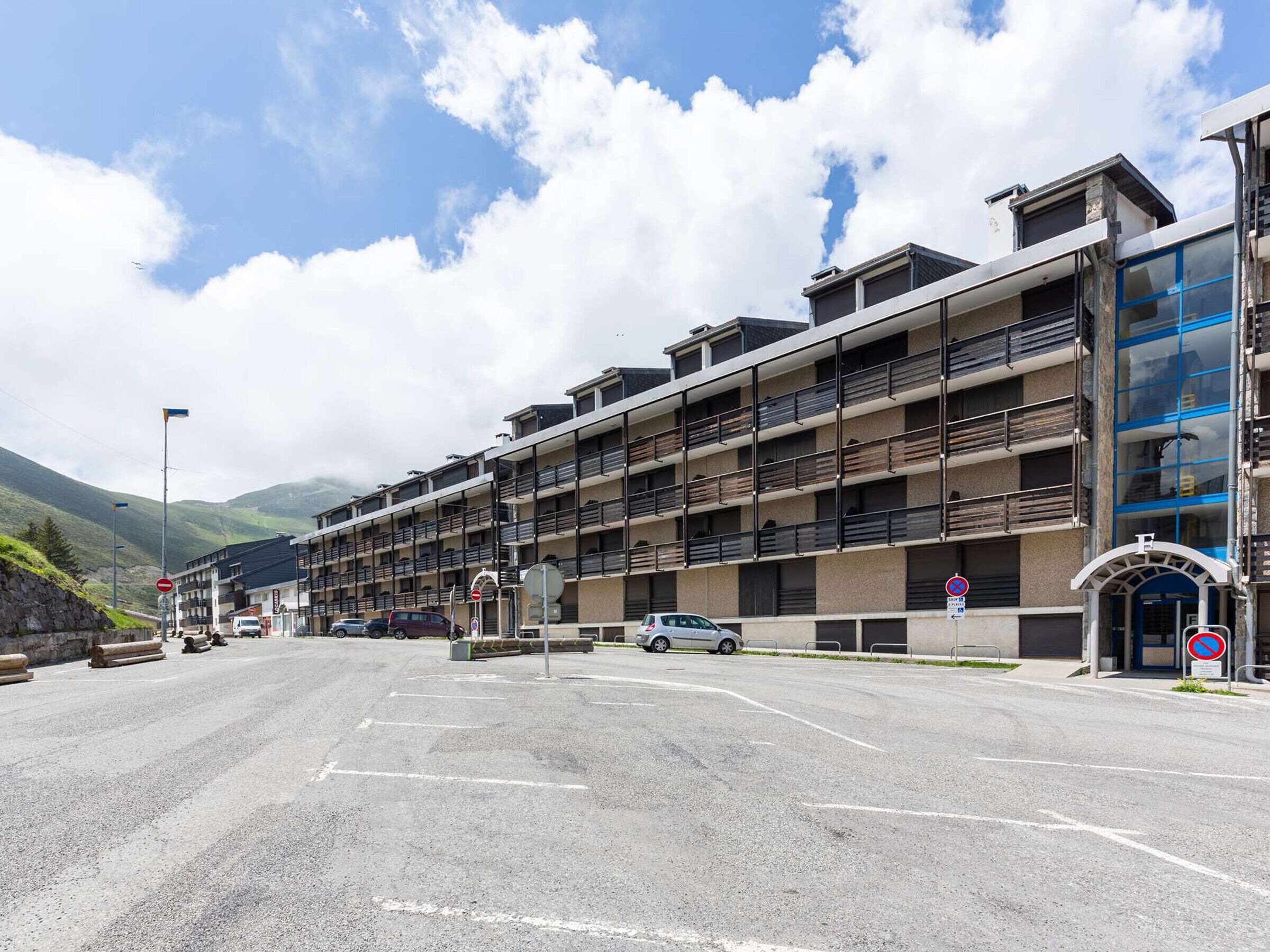 Un hotel de varios pisos con balcones, un estacionamiento y un paisaje montañoso de fondo.