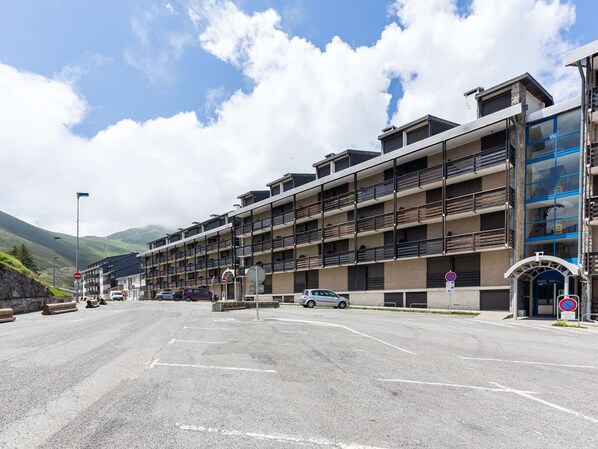 A multi-story hotel with balconies, a parking area, and a mountainous backdrop.