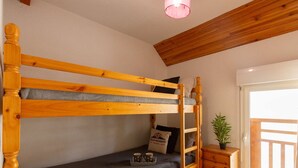 A bunk bed with a grey mattress, a small bedside table with a plant, and a bag labeled "BELLES VACANCES" on the floor.