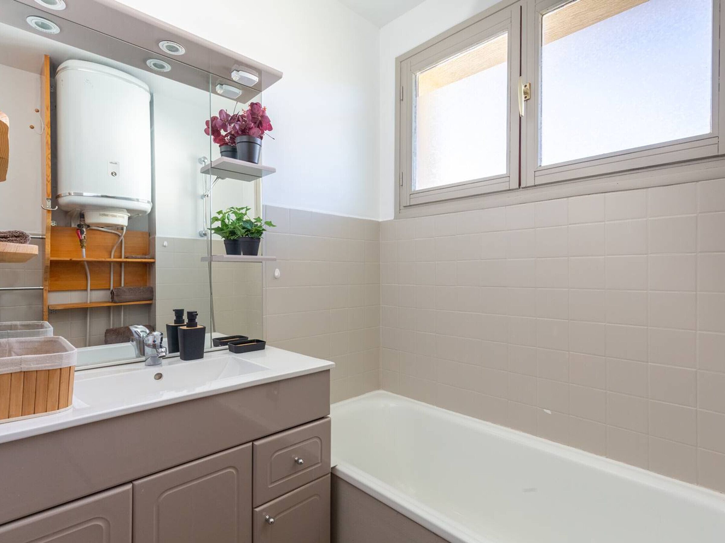 A modern bathroom with a bathtub, a vanity with a mirror, shelves with plants, and a window.