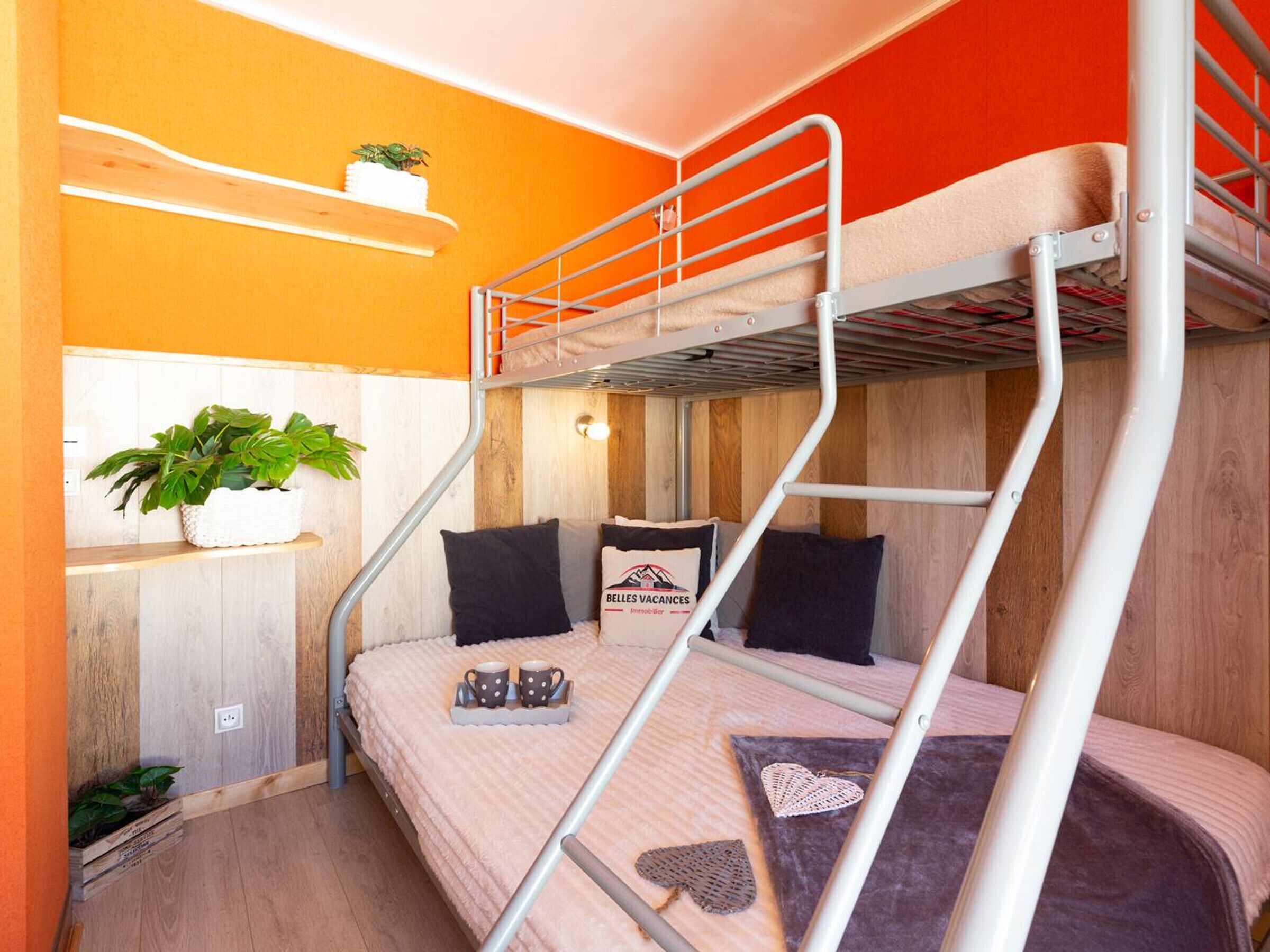 A bunk bed room with a wooden floor, a shelf with a plant, and a pillow with a logo.