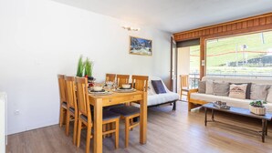 A dining area with a wooden table and chairs, a sofa, a window with a view of a grassy field, and a painting on the wall.