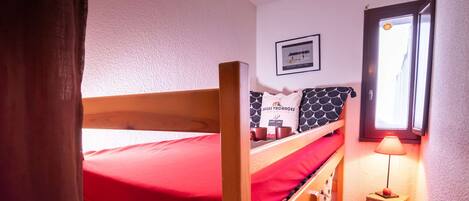 A bunk bed with red bedding, a small bedside table with a lamp, and a window with a view.