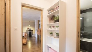 A hallway with a wooden door, a white bookshelf with plants, and a glimpse of a living room with a sofa and a dining area. - Exceptional Duplex 10 pers, Jacuzzi, parking, Saint-Lary (Saint-Lary-Soulan)