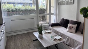 A modern living room with a sofa, a dining table set with plates, a potted plant, and a view through large windows. - Apartment 4 ppl. Saint-Lary, Near center, WIFI (Saint-Lary-Soulan)