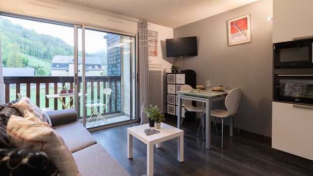 A modern living room with a sofa, a dining table with chairs, a TV mounted on the wall, and a kitchen area with a microwave. - Renovated apartment for 4, quiet, near Saint-Lary center (Saint-Lary-Soulan)