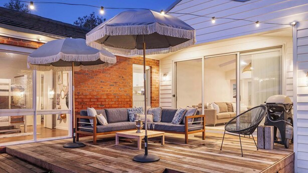 Een terras buiten met houten vloer, twee comfortabele banken, een ronde tafel en twee grote parasols.