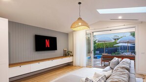 Een moderne woonkamer met een flatscreen televisie, een ruime bank en een glazen deur naar een buitengebied met parasols.
