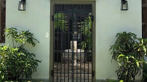 A white building with a black gate, two wall-mounted lights, and a small garden area with plants and a stone pathway. - Samaragiri (Galle)