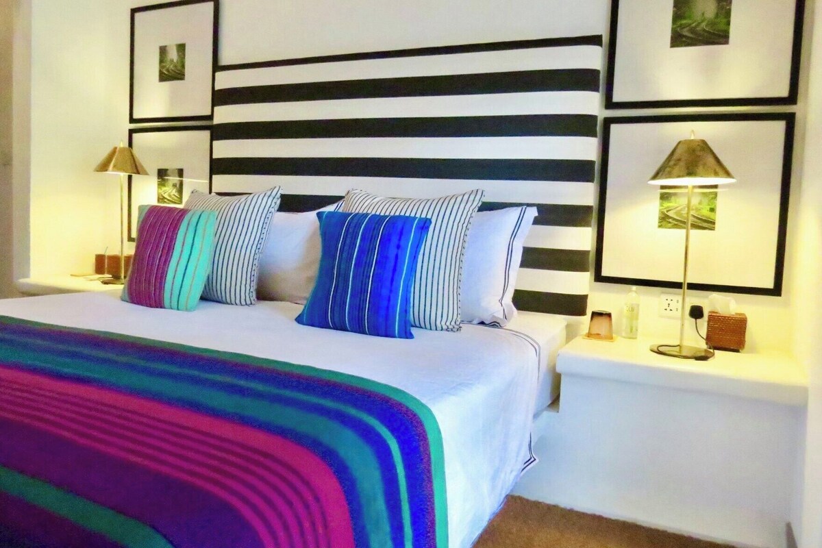 A neatly made bed with striped pillows, a striped headboard, and a colorful blanket. There are two bedside tables with lamps and a small nightstand with a lamp.