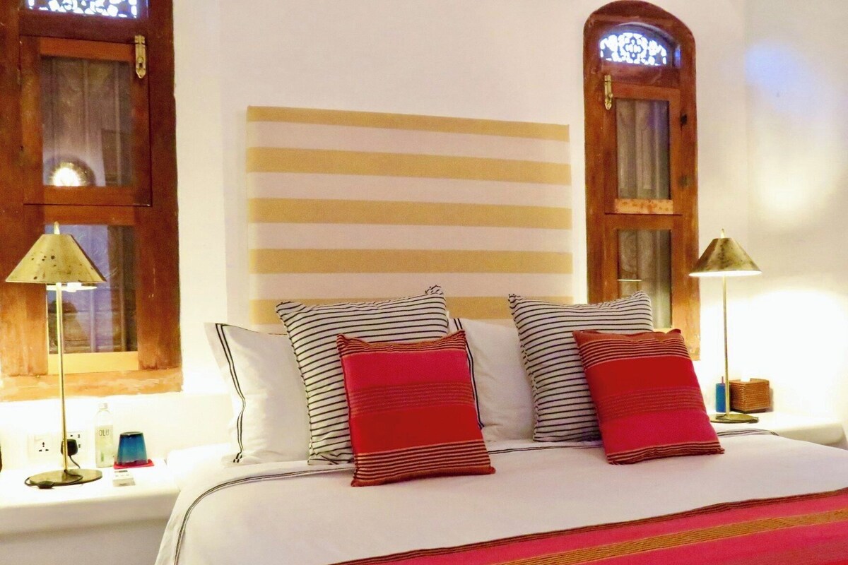 A hotel room with a bed, striped pillows, a red accent pillow, a striped headboard, and a window with a wooden frame.
