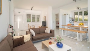Una sala moderna con un sofá marrón, un banco de madera y una mesa de centro blanca. Hay una zona de cocina con una mesa de comedor y bancos, y un televisor en la sala.