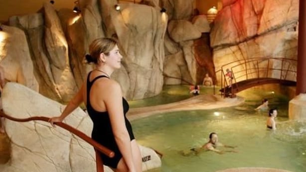 Une femme vêtue d’un maillot de bain noir, debout près d’une piscine dont les parois sont rocheuses et qui est surmontée d’un pont.