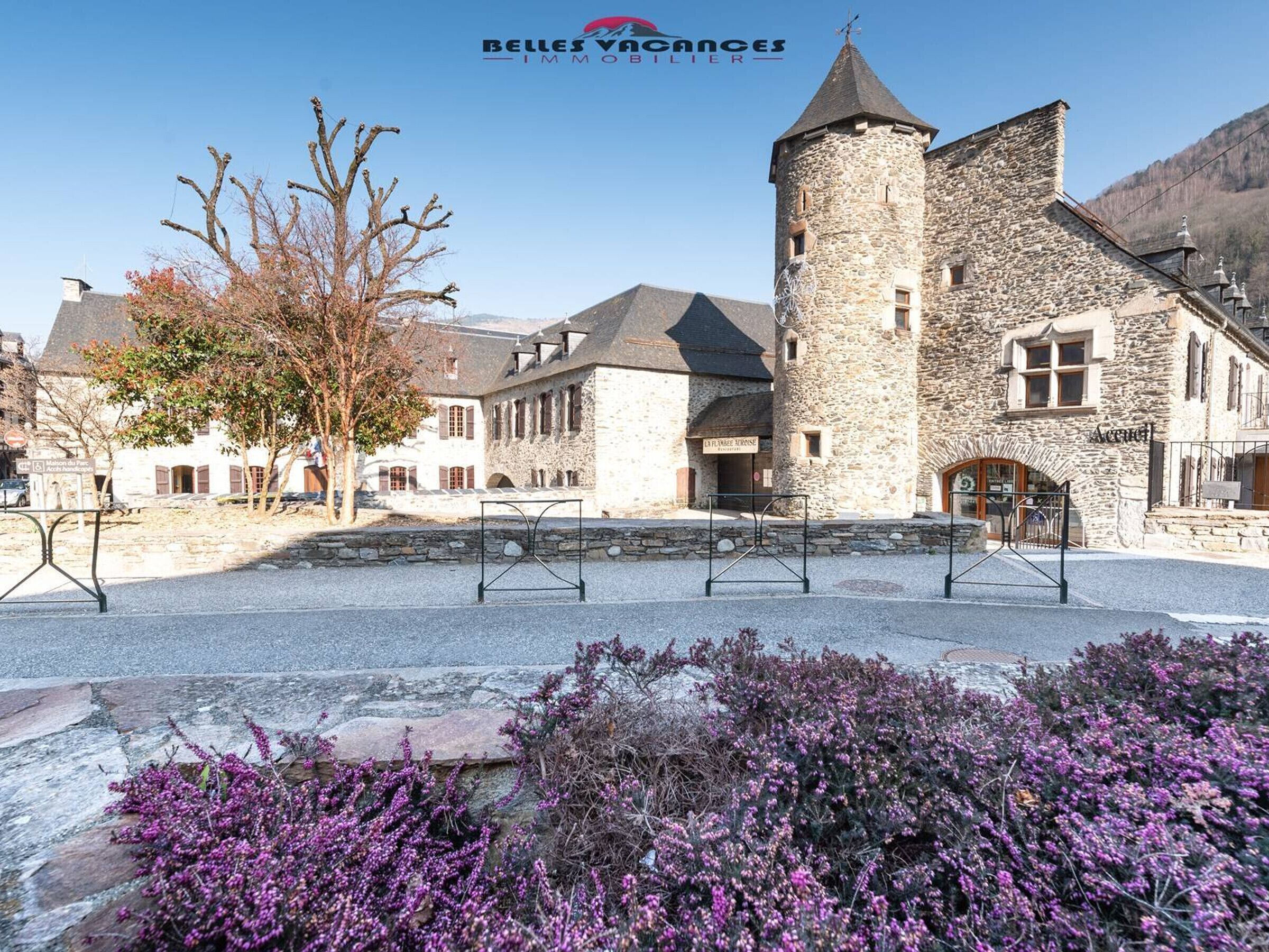 A stone building with a tower, surrounded by a gravel courtyard and purple heather plants.
