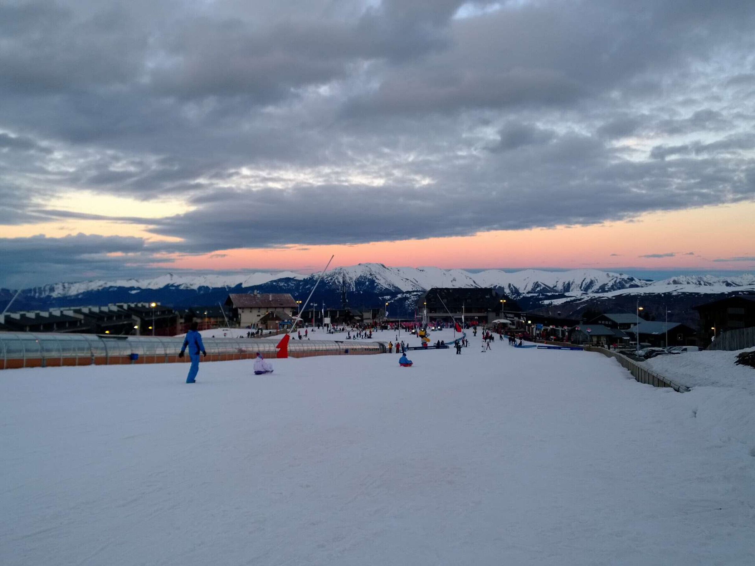 A snowy ski slope with buildings and a mountain range in the background.