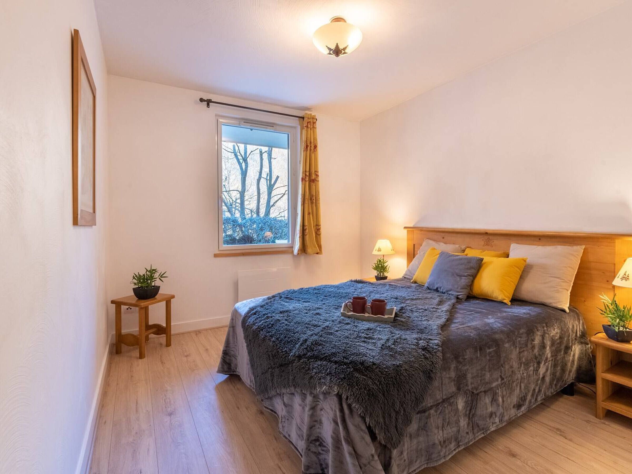 Quarto com cama, janela com cortinas, mesa de cabeceira com abajur e uma pequena planta.