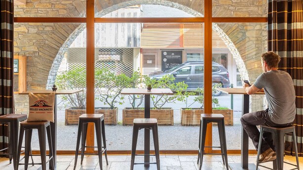 A man sitting at a table inside a cafe with a stone wall and wooden ceiling, overlooking a street with parked cars and a building with a sign. - Duplex Apartment for 8 with Pool and Sauna in City Center (Saint-Lary-Soulan)