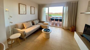 A modern living room with a sofa, a coffee table, and a TV. There is a balcony with a table and chairs.