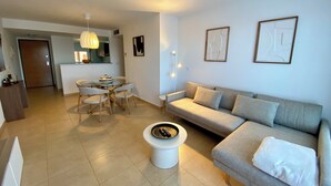 A modern living room with a sofa, a dining table with chairs, a small white table with a decorative item, and a kitchen area in the background.