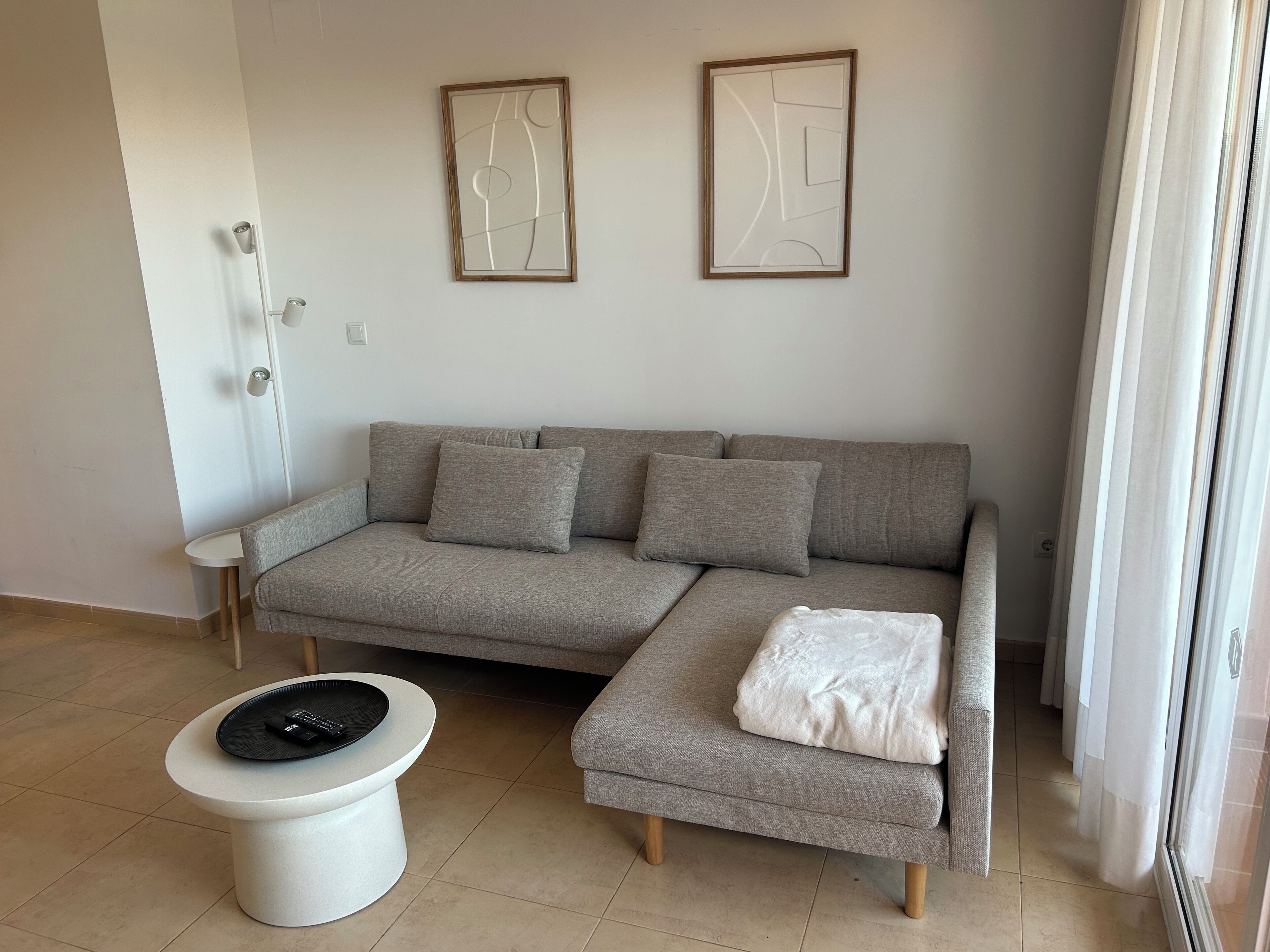 Een moderne woonkamer met een grijze hoekbank, een klein wit bijzettafeltje en een witte poef. Boven de bank hangen abstracte kunstwerken.