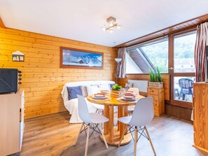 Una habitación acogedora con detalles de madera, una mesa redonda con sillas, una cama con colcha blanca y una ventana con cortinas.
