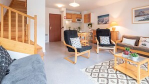 Een ruime woonkamer met een houten trap, twee fauteuils, een bank, een salontafel en een keuken op de achtergrond.