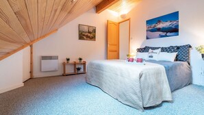 Een slaapkamer met een schuine kap, een bed met beddengoed met een patroon, een wandheater, een klein plankje met planten en een schilderij van een berglandschap.